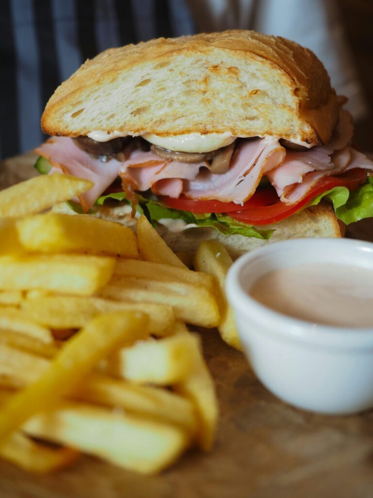 pexels-photo-14693835-14693835 Close-up of a ham sandwich, crispy fries, and a side of sauce on a wooden surface.